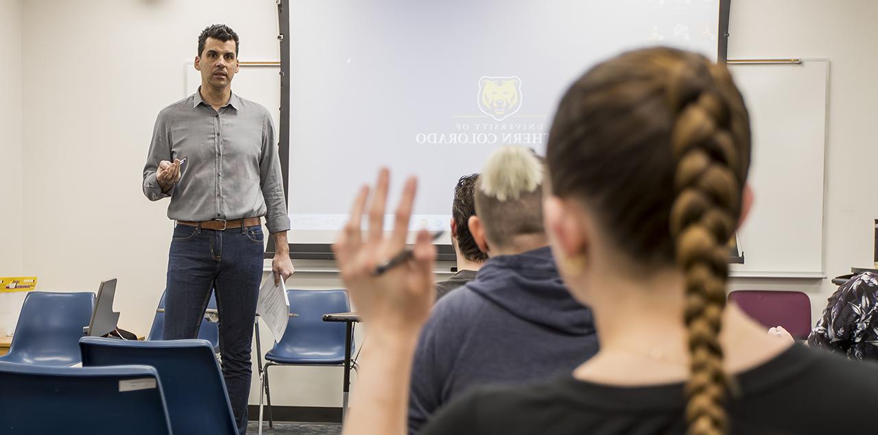 A young college student with their hand up in a classroom with the instructor standing at the front.