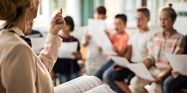 老师站在面前，打开乐谱，对着年幼的学龄儿童唱歌